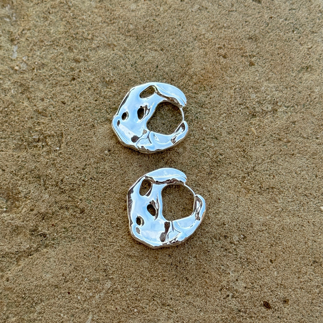 A pair of handmade silver earrings lying on a sandy surface, reflecting Pilgrim's blend of timeless Danish design and trendy style.