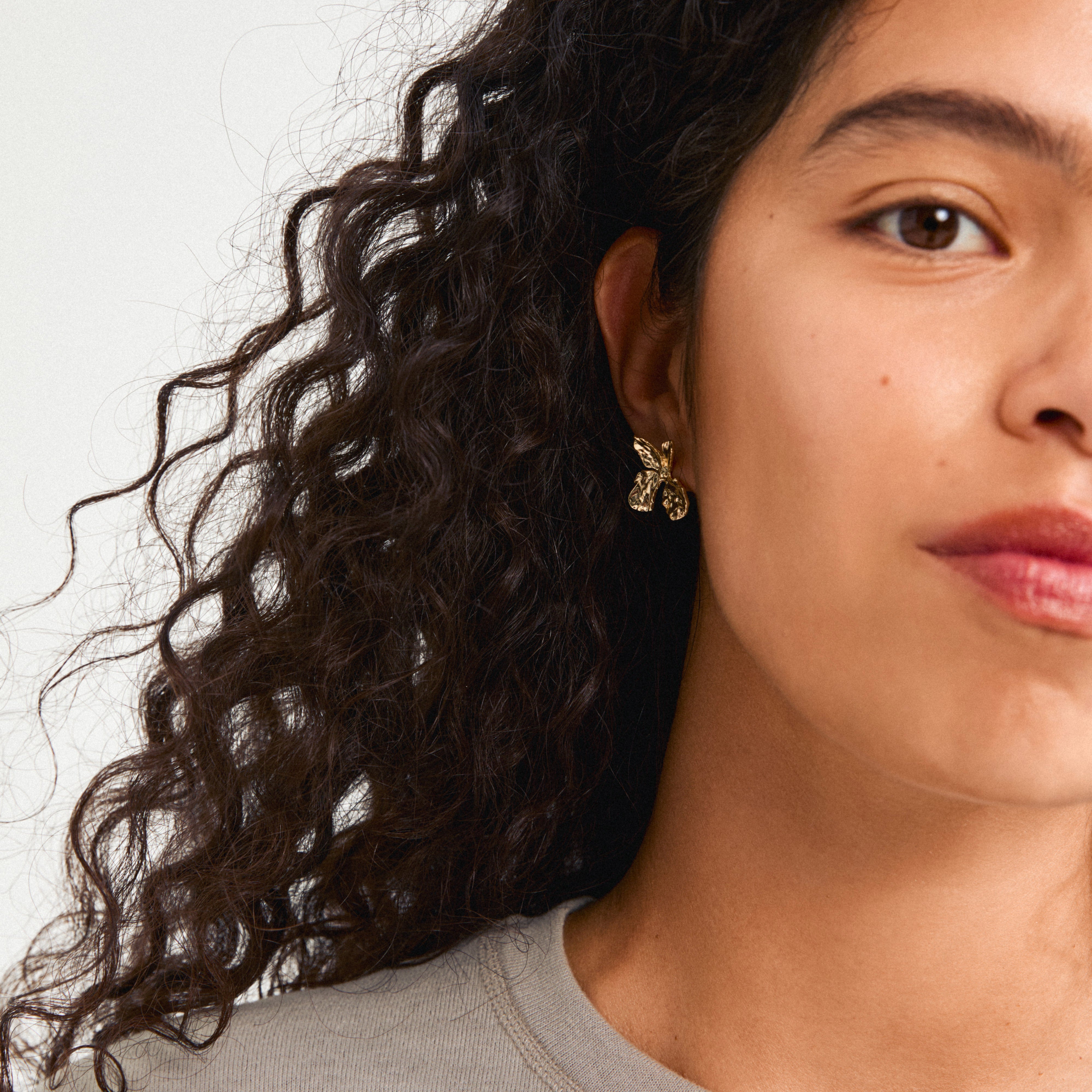 Close-up of Pilgrim's DREAM earrings, featuring gold-plated floral studs inspired by nature, worn by a woman with curly hair.