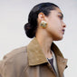 FREJA WEWER x PILGRIM Napoli earrings, gold-plated, showcased on a woman with a bun wearing a coat, highlighting the chunky, square design.