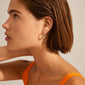 Woman wearing EILEEN twirl hoop earrings, gold-plated with twisted design, complementing her orange tank top and short brown hair.