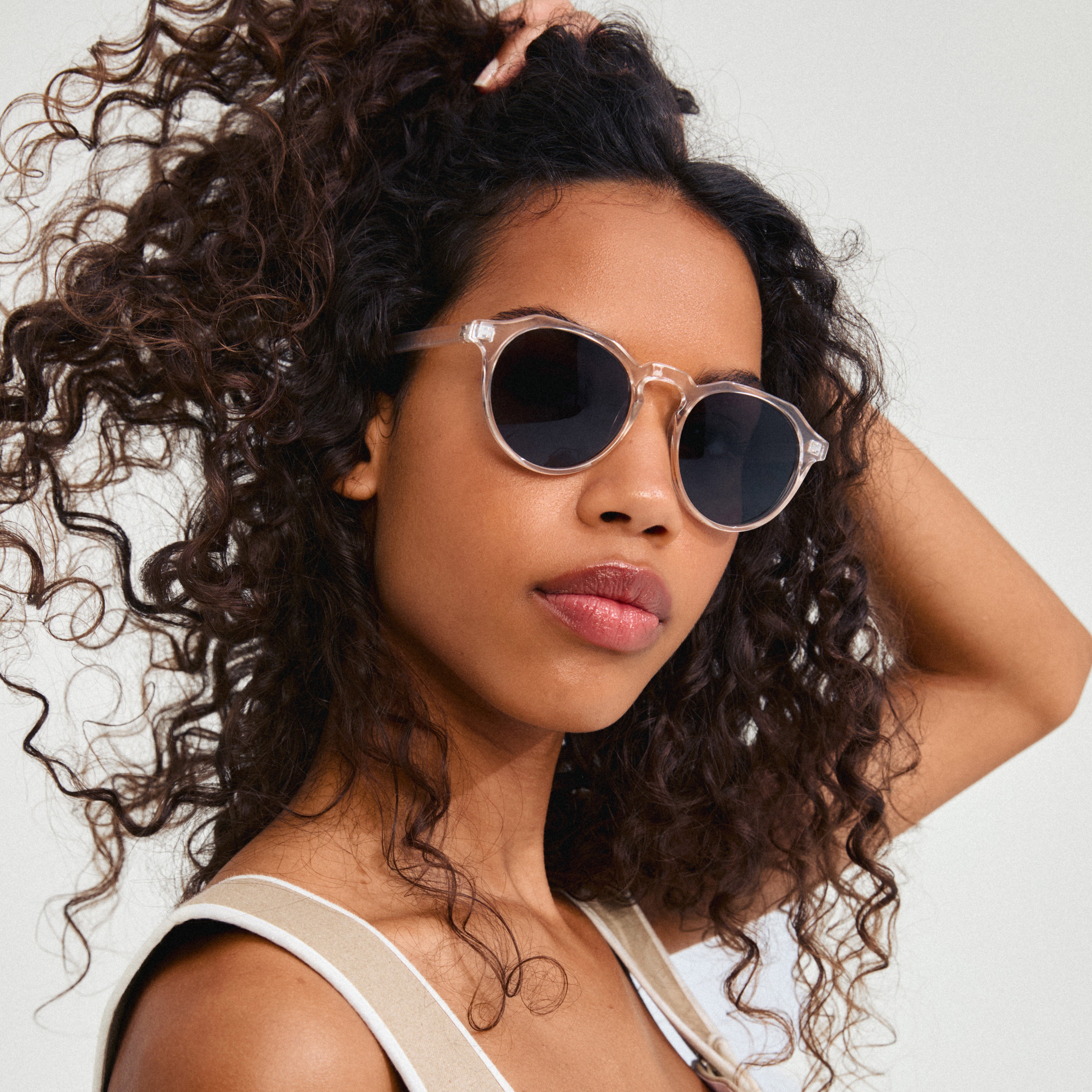 Woman with curly hair wearing ALEJANDRA sunglasses, featuring crystal clear frames and smoke-colored lenses, embodying Pilgrim's trendy, inclusive style.