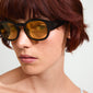 ARIAH sunglasses in black with yellow lenses, worn by a woman with short red hair, featuring super-wide temples and offering UVA/UVB protection.