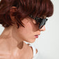 A woman with short red hair wears AUBREE sunglasses with a black frame and crystal accents, embodying a sleek, trendy style from Pilgrim.